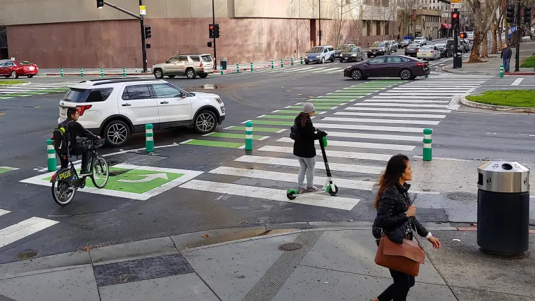 A street intersection with a car, cyclist and scooter in the street, and a pedestrian on the sidewalk.
