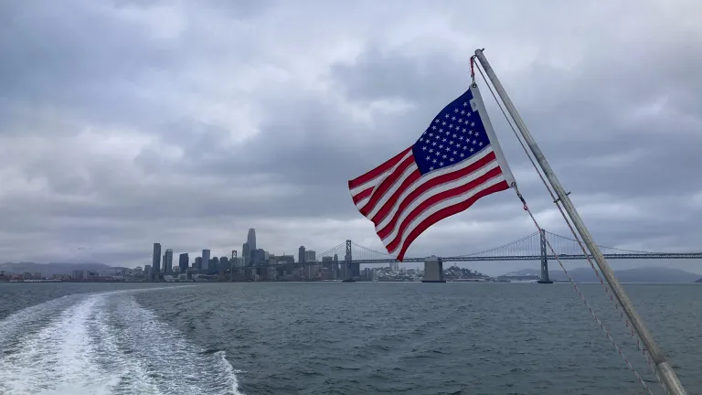 An American flag with the San Francisco-Oakland Bay Bridge and San Francisco skyline in the background.