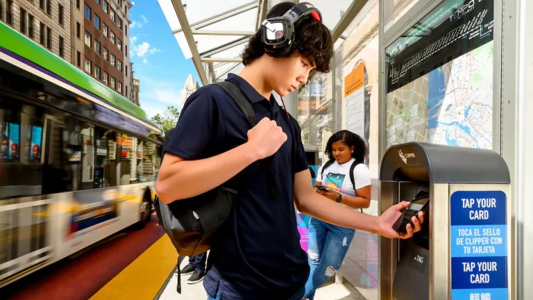 A young man wearing headphones taps his phone on a Clipper reader while an AC Transit bus drives past.