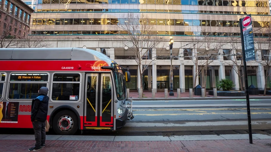 A Muni bus drives on Market Street as a man waits at a bus stop in San Francisco.