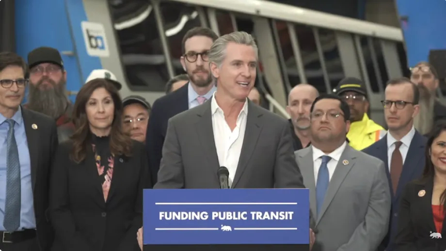Gov. Newsom speaking at a podium with elected officials and BART cars behind him.