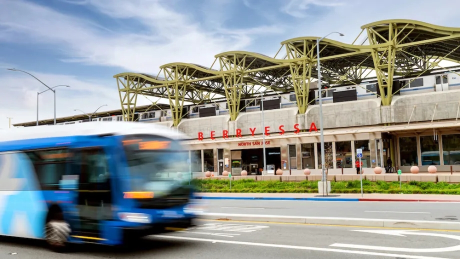 A VTA bus with motion blur drives past Berryessa North San Jose station, with a BART train on the top platform in the background.. 