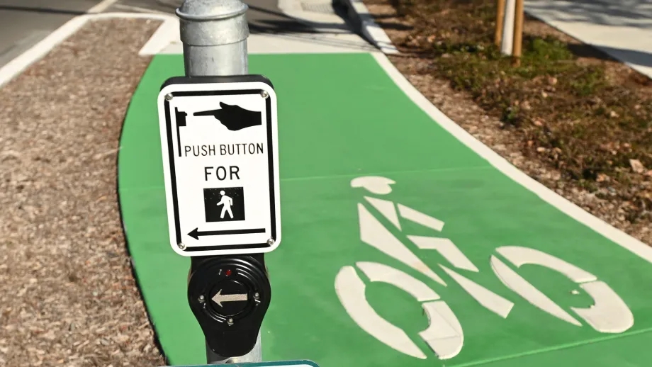 A green separated bike lane and a sign for pedestrians to push a button to cross the street safely.